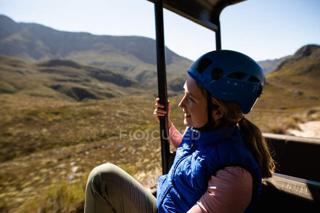 Vista lateral de la mujer caucásica disfrutando del tiempo en la naturaleza, en equipo de forro de cremallera sentado en un coche en un día soleado en las montañas - foto de stock