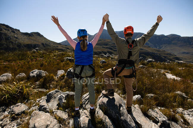 Rear view of Caucasian couple enjoying time in nature together, wearing zip lining equipment, holding hands with arms in the air on a sunny day in mountains — Stock Photo