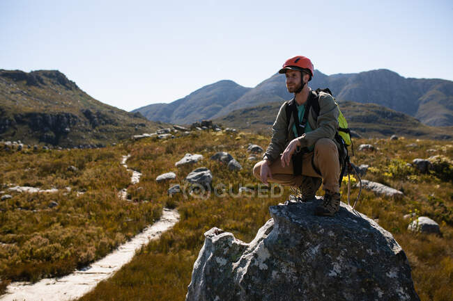 Side view of Caucasian man enjoying time in nature, wearing zip lining equipment, hiking, squatting on a rock on a sunny day in mountains — Stock Photo