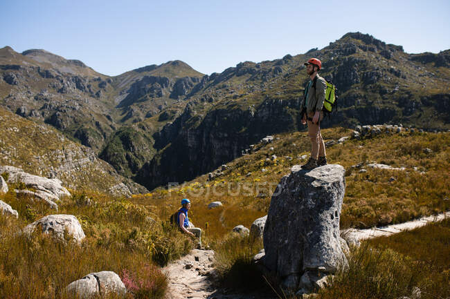Side view of Caucasian couple enjoying time in nature together, wearing zip lining equipment, hiking, the man standing on a rock on a sunny day in mountains — Stock Photo