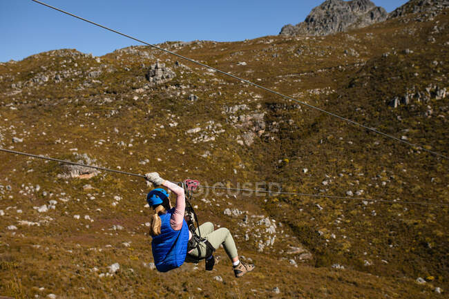 Rear view of Caucasian woman enjoying time in nature, zip lining on a sunny day in mountains. Fun adventure vacation weekend. — Stock Photo