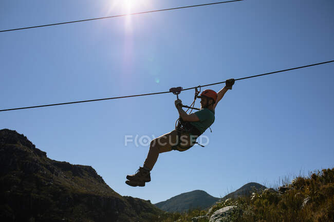 Side view of Caucasian man enjoying time in nature, zip lining on a sunny day in mountains. Fun adventure vacation weekend. — Stock Photo