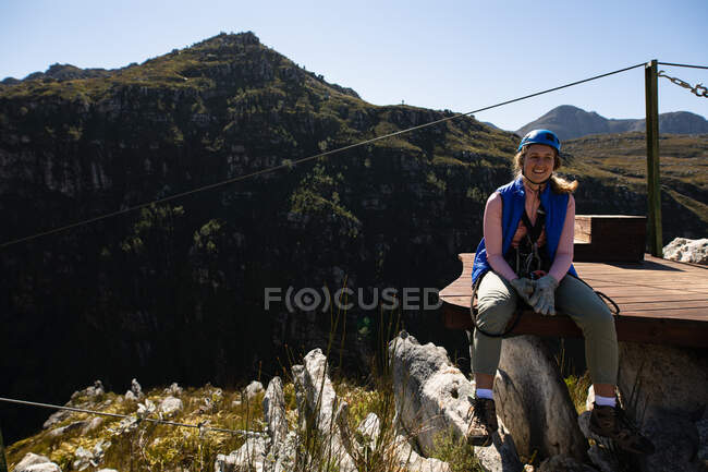 Retrato de mujer caucásica disfrutando del tiempo en la naturaleza, con equipo de forro de cremallera, sentado y sonriendo a la cámara en un día soleado en las montañas - foto de stock