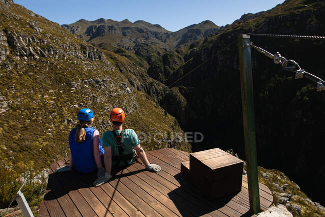 Vue arrière du couple caucasien profitant du temps dans la nature ensemble, portant un équipement de tyrolienne, assis par une journée ensoleillée dans les montagnes — Photo de stock