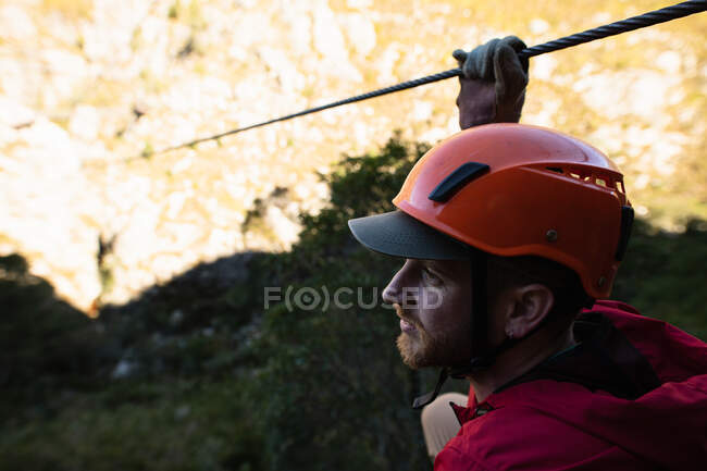 Vista lateral de perto do homem caucasiano desfrutando do tempo na natureza, vestindo equipamento de tirolesa, segurando corda de tirolesa em um dia ensolarado nas montanhas — Fotografia de Stock