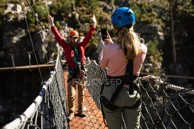 Rear view of Caucasian couple enjoying time in nature, wearing zip lining equipment, the woman taking pictures of a man with a smartphone on a sunny day in mountains — Stock Photo