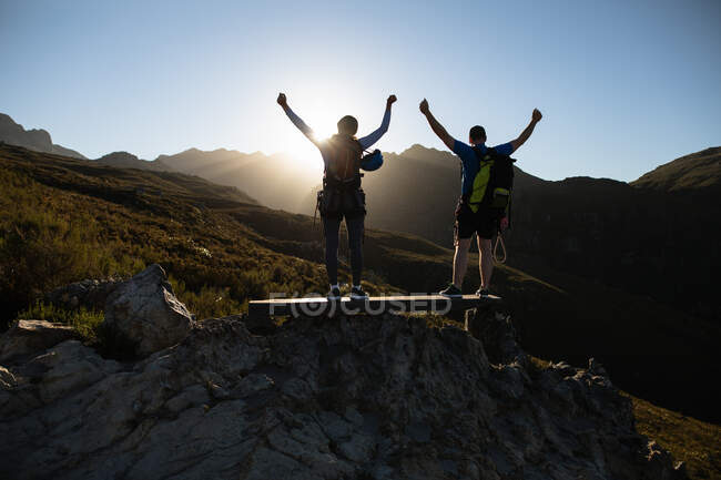 Rear view of Caucasian couple enjoying time in nature together, wearing zip lining equipment, hiking, standing on a rock, arms in the air, on a sunny day in mountains — Stock Photo