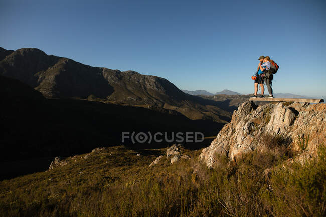 Vista laterale della coppia caucasica godendo del tempo nella natura insieme, indossando attrezzature zip fodera, escursioni, in piedi su una roccia, abbracci, in una giornata di sole in montagna — Foto stock
