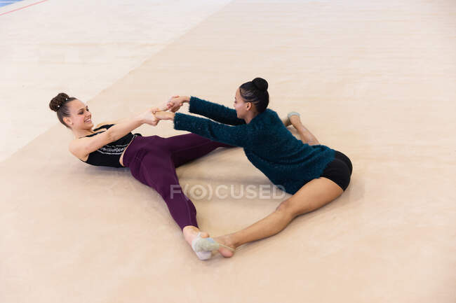 Vista lateral de ángulo alto de gimnastas femeninas de raza caucásica y mixta que practican juntos en el gimnasio, sentados en el suelo y estirándose - foto de stock