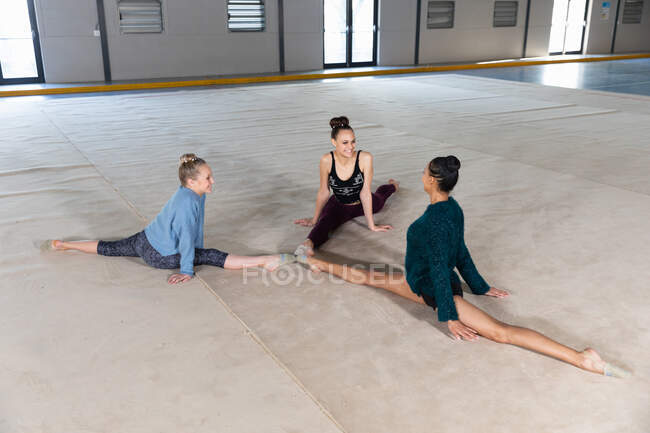 Vista de ángulo alto de una raza mixta adolescente y dos gimnastas femeninas caucásicas practicando juntos en el gimnasio, haciendo splits, tocándose los dedos de los pies, estirándose y sonriéndose - foto de stock