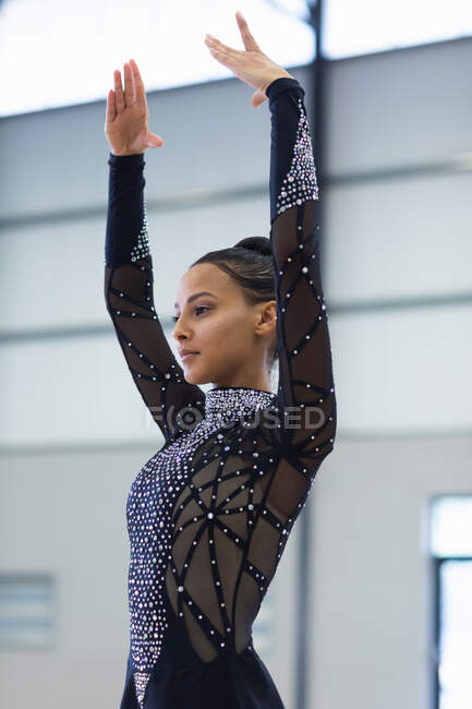 Вид сбоку на юношескую гимнастку смешанной расы, выступающую в тренажерном зале, стоящую с поднятыми руками, в черном трико — стоковое фото