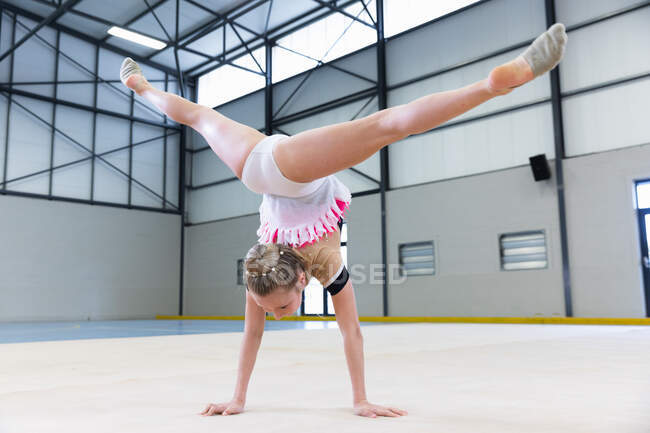 Вид сзади на юную белую, розовую и бежевую гимнастку, выступающую в спортзале, делающую стойку на руках и разделяющуюся, в белом, розовом и бежевом трико — стоковое фото
