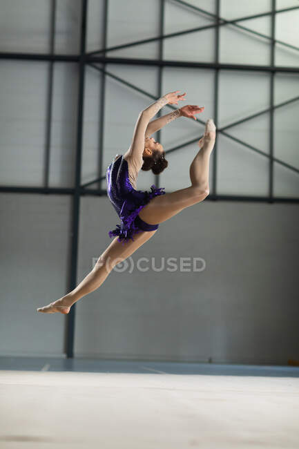 Vista laterale della ginnasta caucasica adolescente che si esibisce in palestra, saltando con le braccia tese, indossando un body viola. — Foto stock