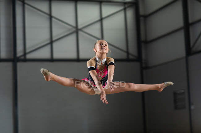 Вид спереди на юную белую гимнастку, выступающую в тренажерном зале, прыгающую и делающую сплит, одетую в розовый и бежевый трико — стоковое фото