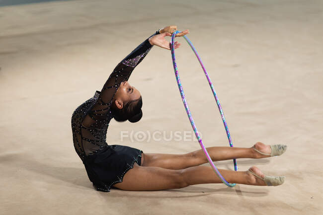 Vue latérale d'une gymnaste féminine de race mixte adolescente jouant au gymnase, faisant de l'exercice avec un cerceau, se penchant vers l'arrière, portant un justaucorps noir — Photo de stock