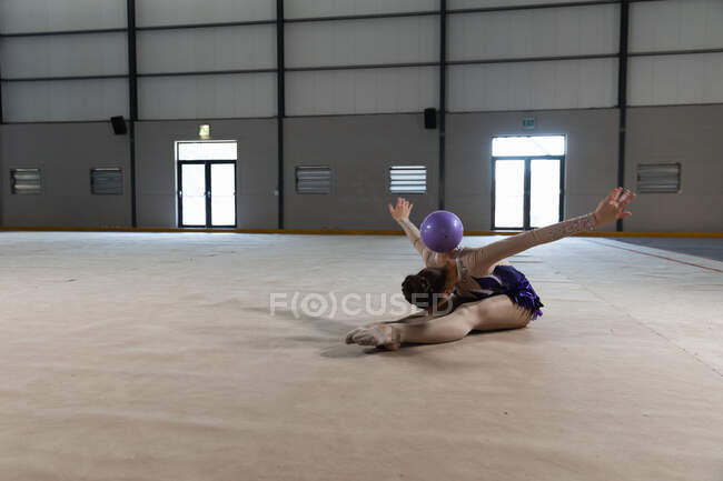 Vista lateral de la adolescente gimnasta caucásica que actúa en el gimnasio, haciendo ejercicio con bola púrpura, sentada en el suelo, la pelota descansando sobre su espalda, usando un maillot púrpura - foto de stock