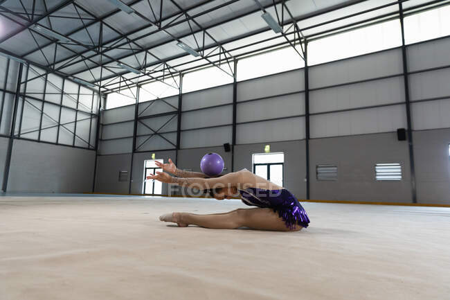 Вид сбоку на юную кавказскую гимнастку, выступающую в спортзале, упражняющуюся с фиолетовым мячом, сидящую на полу с вытянутыми руками, шар, лежащий на спине, в фиолетовом трико — стоковое фото