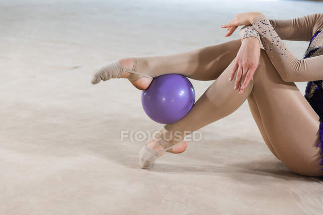Vista laterale bassa sezione della ginnasta femminile esibendosi in palestra, esercitandosi con palla viola, seduto sul pavimento, la palla tenuta per le gambe, indossando body viola — Foto stock