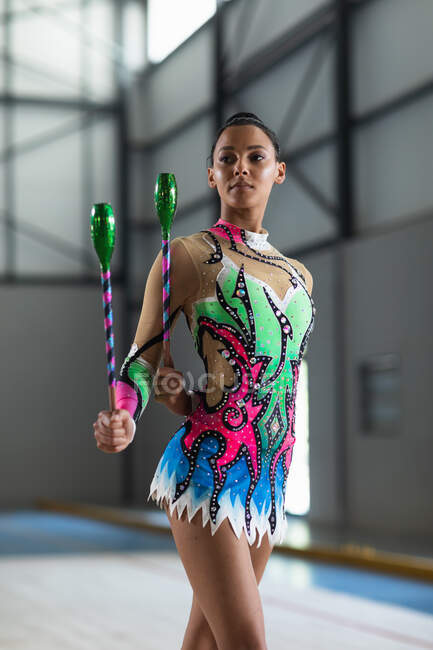 Front view of teenage mixed race female gymnast performing at the gym, exercising with clubs, holding clubs, concentrating, wearing multi colored leotard — Stock Photo