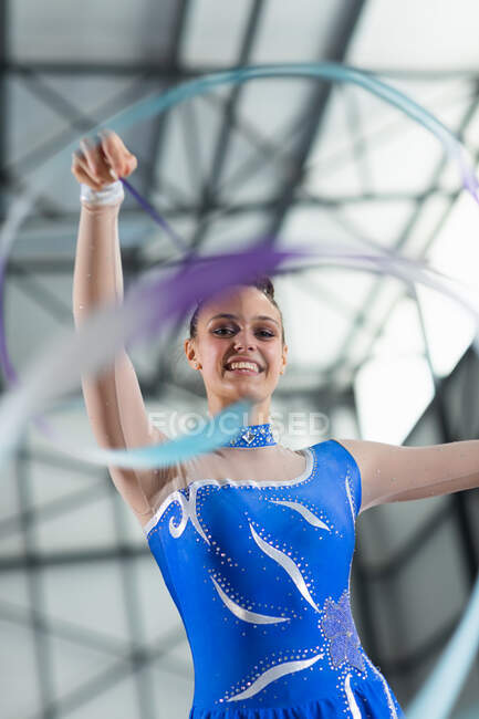 Портрет підлітка-кавказької гімнастки, що виступає в спортзалі з стрічкою, одна рука, витягнута, дивлячись на камеру, одягнена в синій трикотажний одяг. — стокове фото