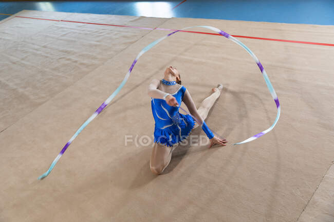 Vista frontal de ángulo alto de la gimnasta femenina caucásica adolescente que actúa en el gimnasio, haciendo ejercicio con cinta, sentado en el suelo, doblándose hacia atrás, usando maillot azul - foto de stock