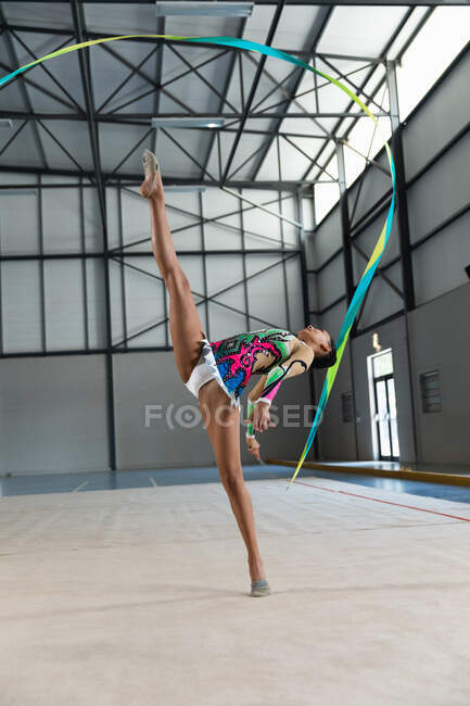 Vista lateral de la gimnasta femenina adolescente de raza mixta que actúa en el gimnasio, haciendo ejercicio con cinta, de pie en split, con maillot multicolor - foto de stock