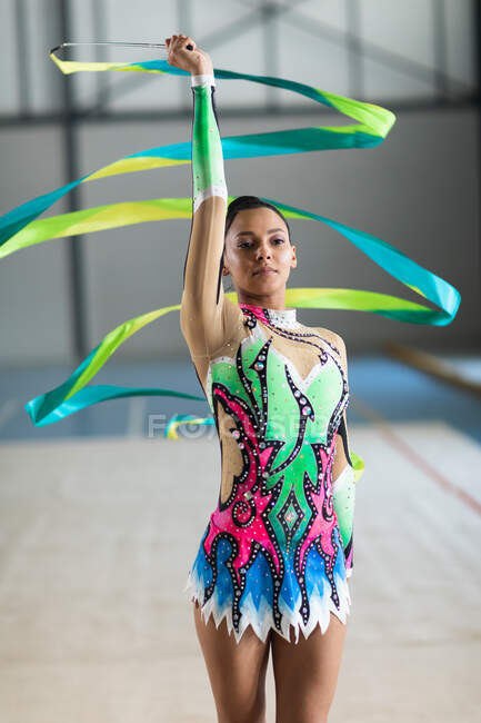 Vista frontal de cerca de la gimnasta femenina adolescente de raza mixta que actúa en el gimnasio, haciendo ejercicio con cinta, usando maillot multicolor - foto de stock