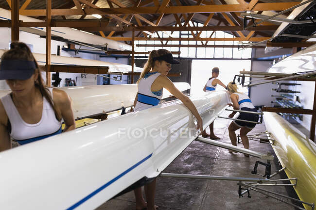 Vue latérale d'une équipe d'aviron de quatre femmes caucasiennes soulevant un bateau dans un hangar à bateaux et se préparant à le porter à la rivière — Photo de stock
