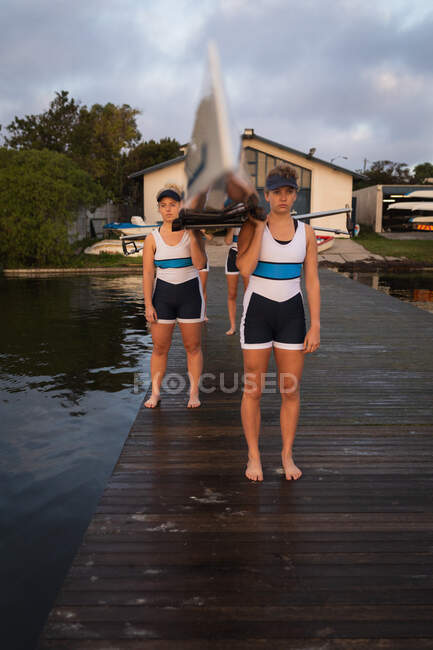 Front view of a rowing team of four Caucasian women carrying a boat on their shoulders, standing on a jetty in the river, with a boathouse in the background — Stock Photo
