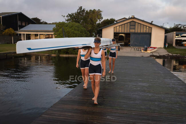 Front view of a rowing team of four Caucasian women carrying a boat on their shoulders, walking along a jetty on the river, with a boathouse in the background — Stock Photo