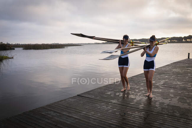 Front view of two Caucasian women from a rowing team carrying oars on their shoulders and walking along a jetty on the river at sunrise, talking and smiling - foto de stock
