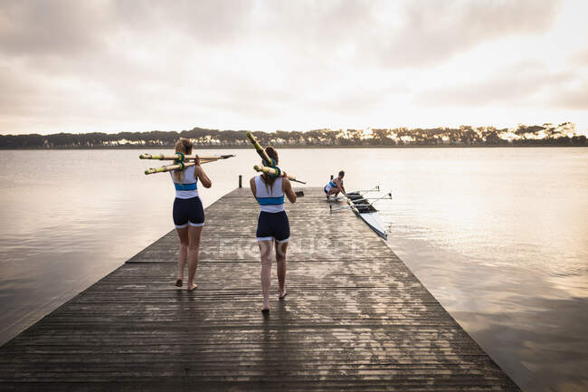 Rear view of two Caucasian women from a rowing team carrying oars on their shoulders and walking along a jetty on the river at sunrise, their teammates preparing a boat on the river in the background — Stock Photo