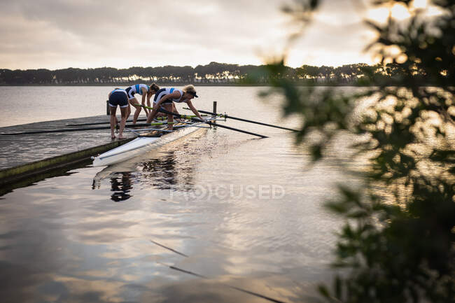 Side view of a rowing team of four Caucasian women standing on a jetty preparing a boat on the river for rowing training, with foliage in the foreground — Stock Photo