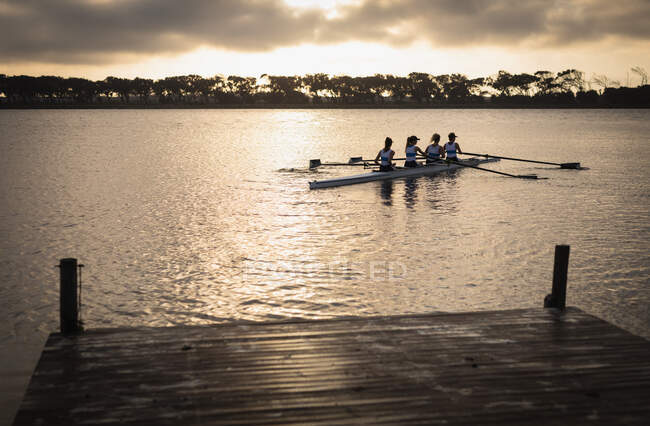 Rear view of a rowing team of four Caucasian women training on the river, rowing in a racing shell at sunrise, with sunlight reflected in the ripples of the water and a jetty in the foreground — Stock Photo