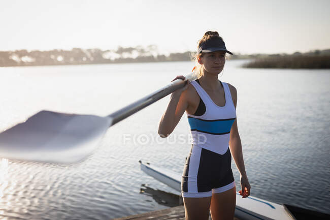 Side view close up of a Caucasian female rower training on the river, standing on a jetty, holding an oar on her shoulder and looking away — Stock Photo