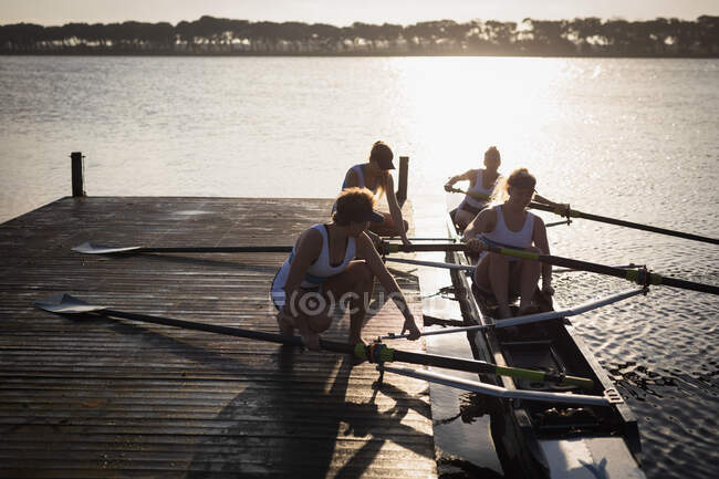 Передній вид веслувального колективу з чотирьох кавказьких жінок тренуватися на річці, на пристані, що готує гоночний снаряд для веслування на світанку. — стокове фото