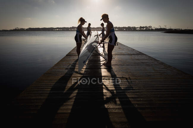Side view of a rowing team of four Caucasian women training on the river, carrying a boat standing on a jetty at sunrise — Stock Photo