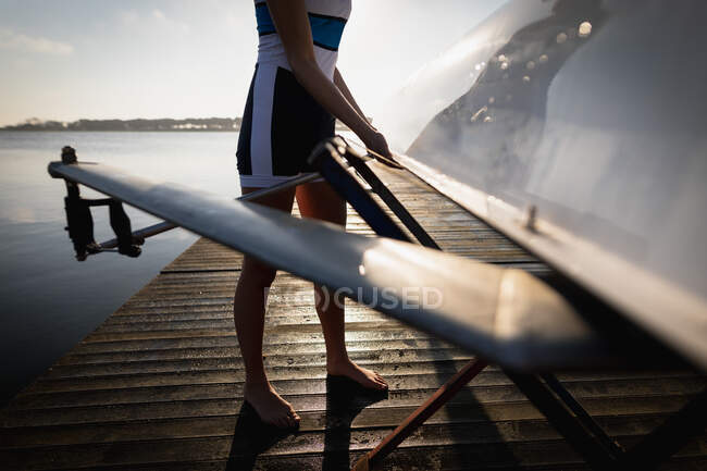 Vista lateral de sección baja de un miembro de un equipo de remo de mujeres entrenando en el río, llevando un barco, de pie descalzo en un embarcadero al amanecer - foto de stock