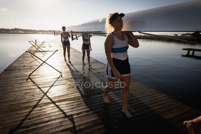 Vista lateral de uma equipe de remo de quatro mulheres caucasianas treinando no rio, carregando um barco em seus ombros andando em um molhe ao nascer do sol — Fotografia de Stock