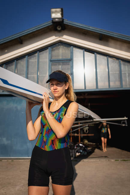 Front view close up of a Caucasian female rower carrying a boat on her shoulder out of a boathouse in the sun, her teammate carrying the other end in the background — Stock Photo