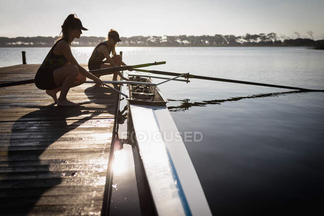 Вид сбоку на двух кавказских гребцов из гребной команды, тренирующихся на реке, стоящих на коленях на причале и готовящих весла на лодке на воде — стоковое фото
