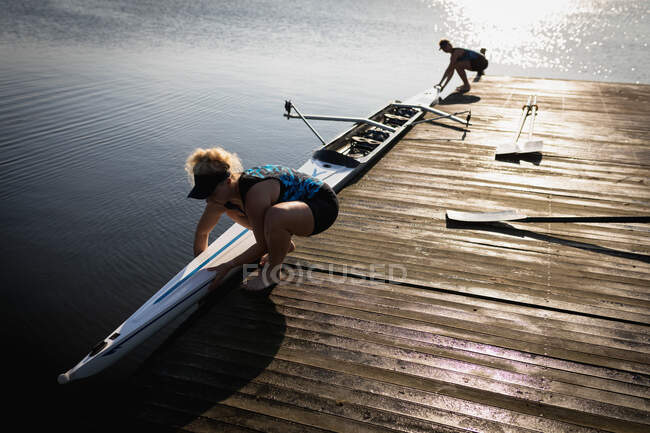 Seitenansicht von zwei kaukasischen Ruderinnen eines Ruderteams, die auf dem Fluss trainieren, auf einem Steg stehen und ein Boot ins Wasser lassen — Stockfoto
