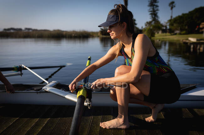 Side view close up of a Caucasian female rower from a rowing team training on the river, kneeling on a jetty and preparing the oar on a boat on the water in the sun — Stock Photo