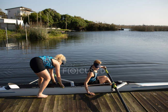 Vista lateral de dos remeros femeninos caucásicos desde un equipo de remo entrenando en el río, subiendo a una concha de carreras en el agua y empujando desde el embarcadero en el sol - foto de stock