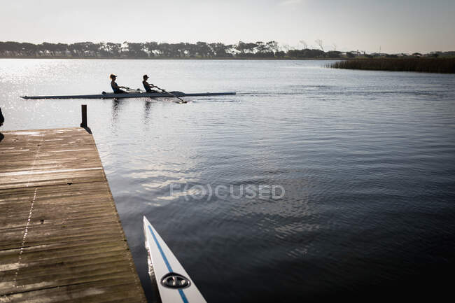 Distant side view of two Caucasian female rowers from a rowing team training on the river, rowing in a racing shell on the water, seen from a jetty — Stock Photo