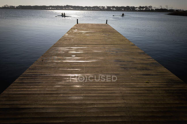 Frontansicht eines Stegs an einem Fluss in der Morgensonne, im Hintergrund Ruderinnen eines Ruderteams, die in zwei Rennmuscheln auf dem Wasser trainieren — Stockfoto