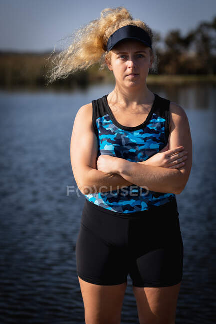 Portrait of a Caucasian female rower from a rowing team with long blonde, curly hair, wearing a visor, standing in the sun with her arms crossed, looking to camera, with the river in the background — Stock Photo