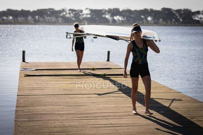 Vorderansicht zweier kaukasischer Ruderinnen von einem Ruderteam, die auf dem Fluss trainieren und ein Boot auf ihren Schultern tragen, das auf einem Steg in der Sonne spaziert — Stockfoto