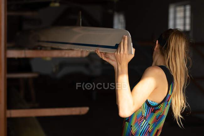 Rear view close up of a Caucasian female rower from a rowing team lifting one end of a boat from a storage rack in a boathouse on her shoulder in the sun — Stock Photo
