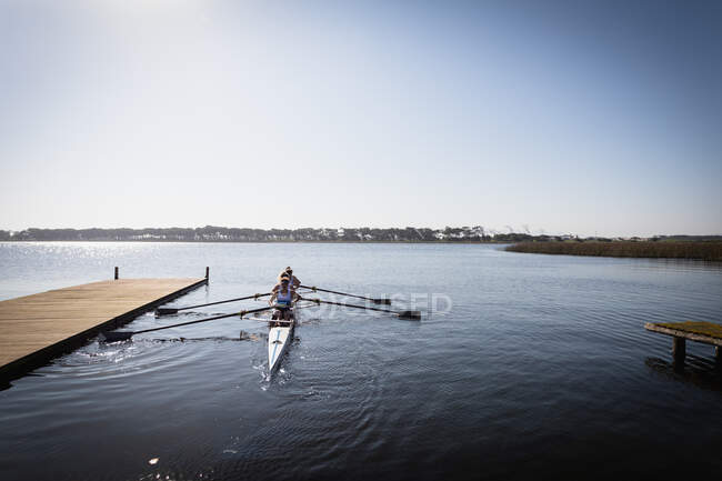 Front view of a rowing team of four Caucasian women training on the river, rowing in a racing shell beside a jetty in the sun — Stock Photo
